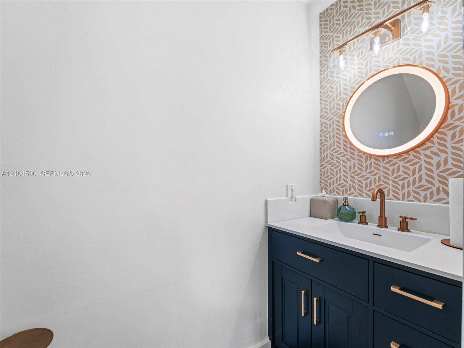 Navy blue vanity with metallic hardware, integrated white countertop, oval LED-backlit mirror, patterned accent wall, and metallic multi-light fixture