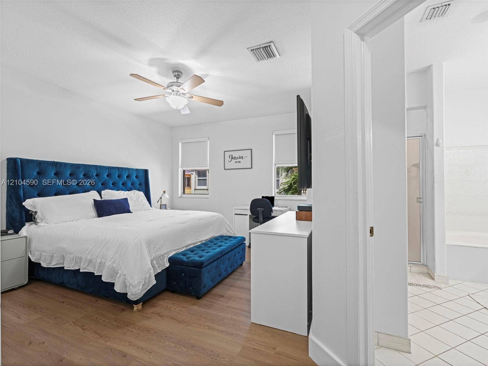 Bedroom featuring wood-finish flooring, a multi-blade ceiling fan with integrated lighting, dual windows, and an en-suite bathroom with tile flooring