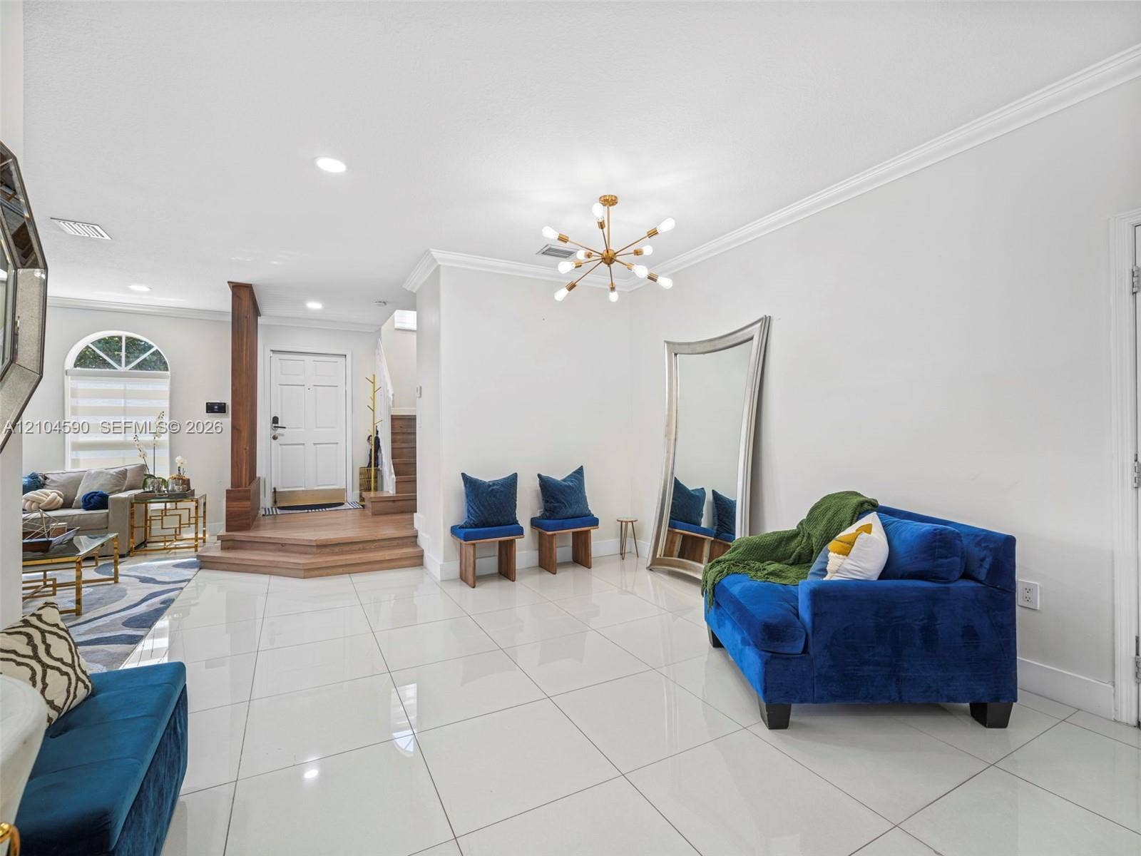 Open-concept interior featuring large-format tile flooring, crown molding, a multi-arm sputnik-style chandelier, and a raised entryway platform with wood-finish flooring