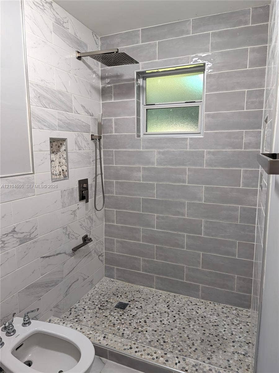 Walk-in shower featuring gray subway-style wall tile, contrasting white stone-finish wall accents, a pebble-finish floor, built-in recessed niche, and metallic rainfall showerhead with a separate hand-held sprayer