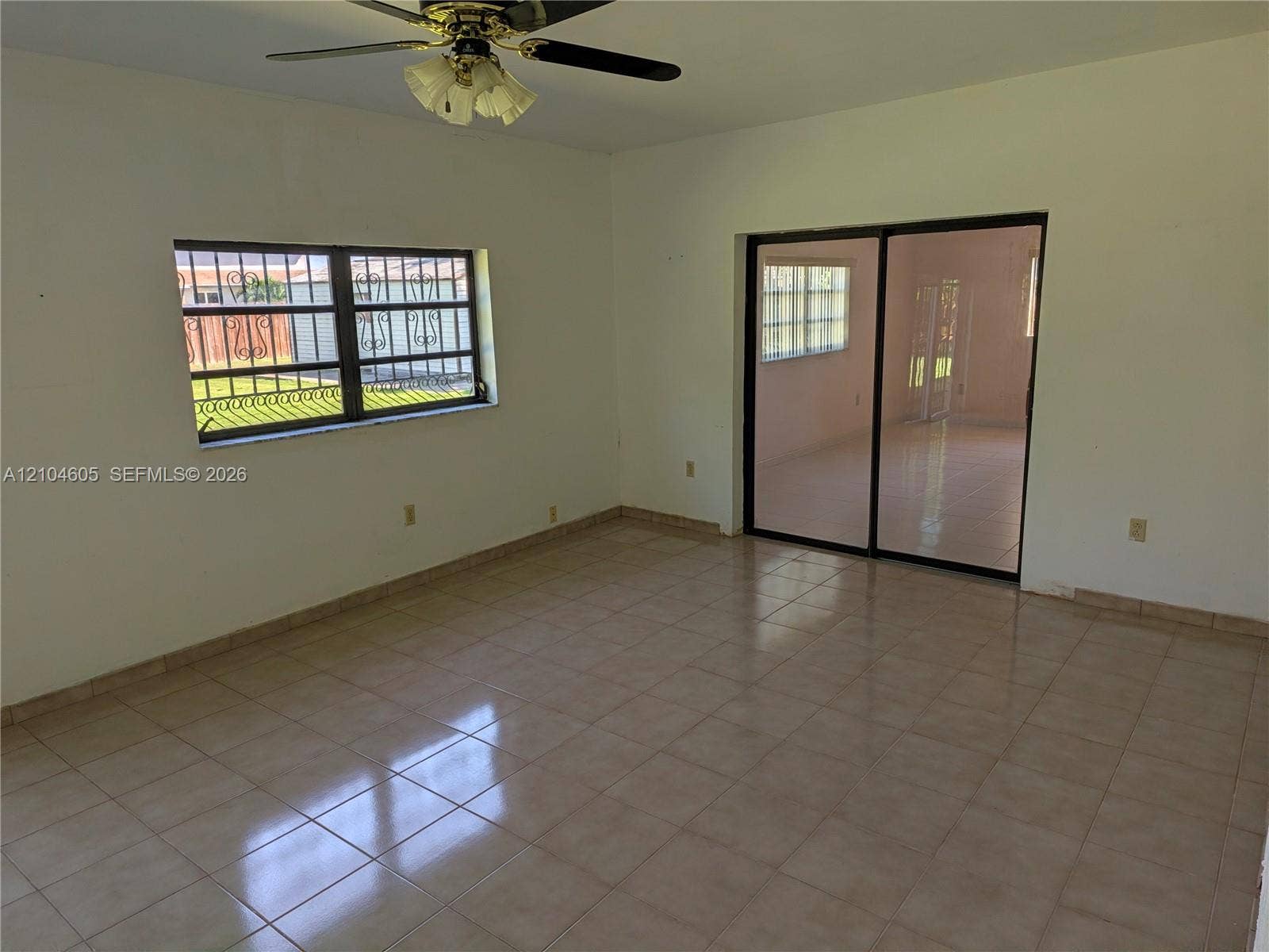 Neutral ceramic tile flooring, sliding glass doors, window with decorative security bars, and a ceiling fan with integrated light fixture
