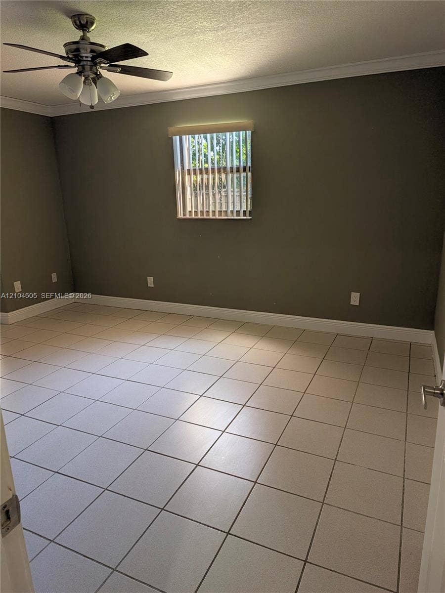 Neutral-toned interior space featuring light-colored square tile flooring, crown molding, a central ceiling fan with multi-light fixture, and a window with vertical blinds
