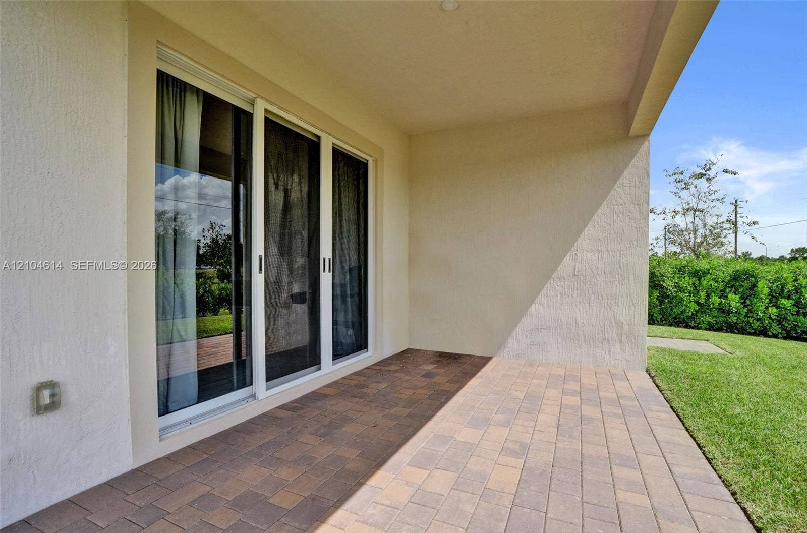 12240 SW 44th Court, Davie, FL 33330 Covered patio with multi-tone paver flooring, sliding glass doors, and a surrounding lawn with perimeter hedging