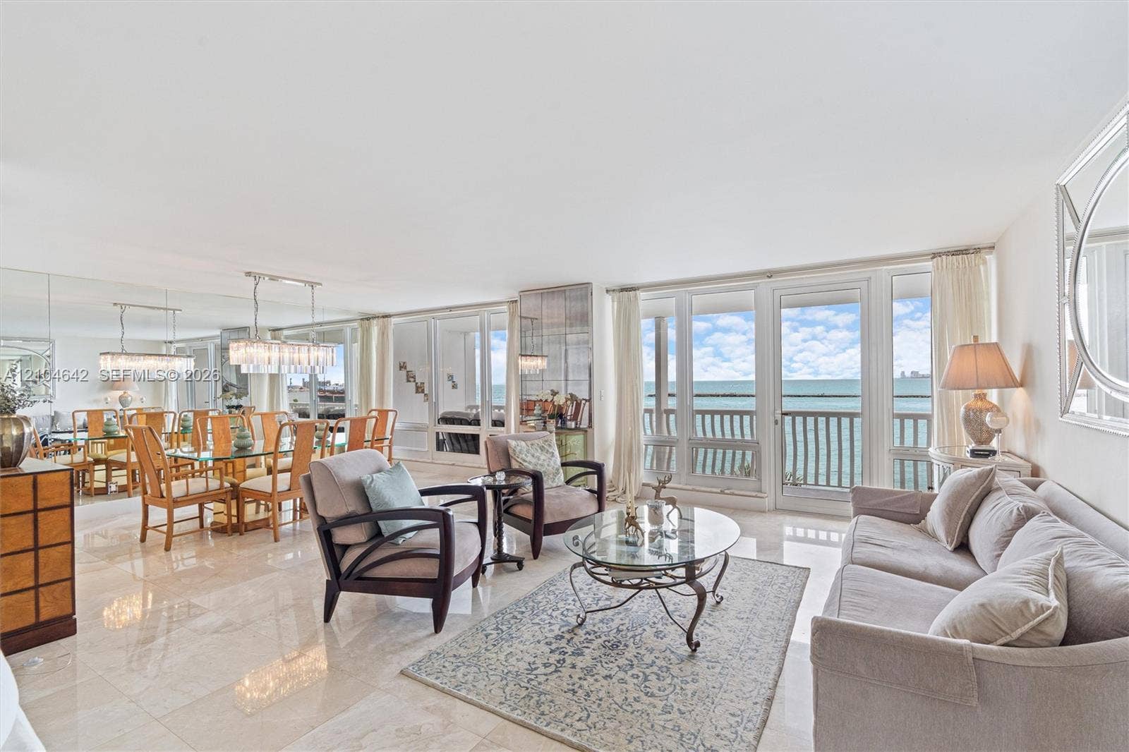 Open-concept living space featuring polished stone flooring, floor-to-ceiling glass paneling, a full-wall mirror, and a balcony with direct water exposure