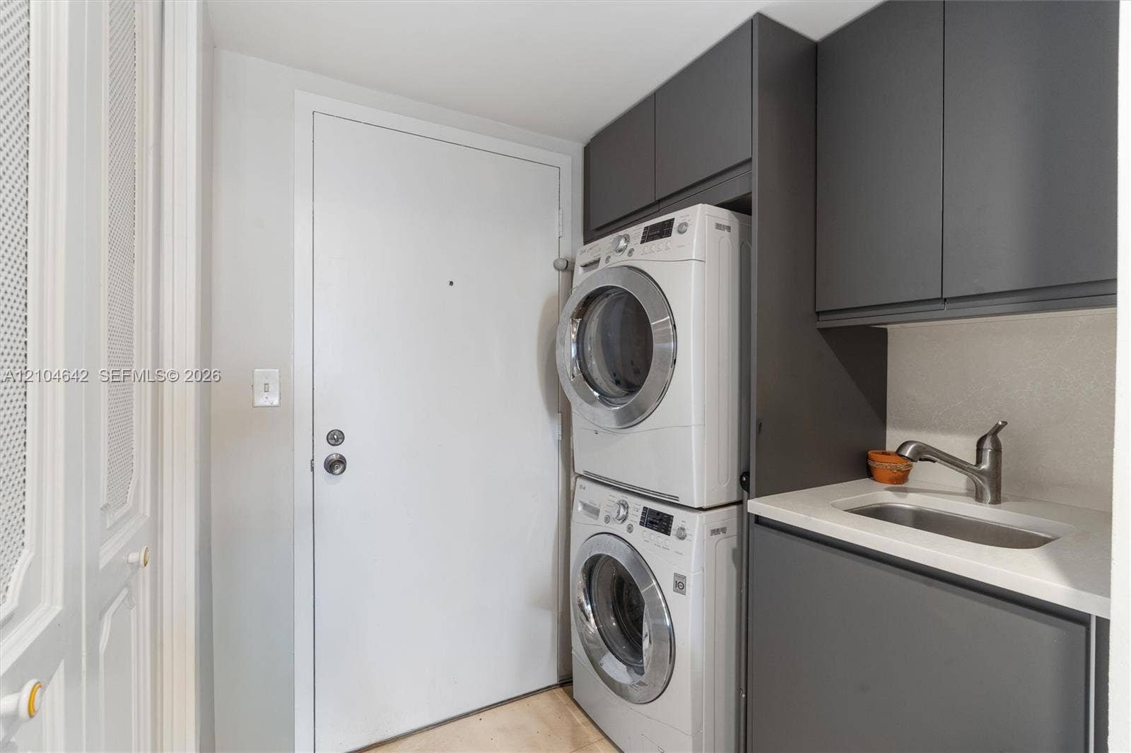 Laundry area featuring charcoal-toned cabinetry, light-toned stone-finish countertop, integrated sink with a gooseneck faucet, and stacked appliance unit