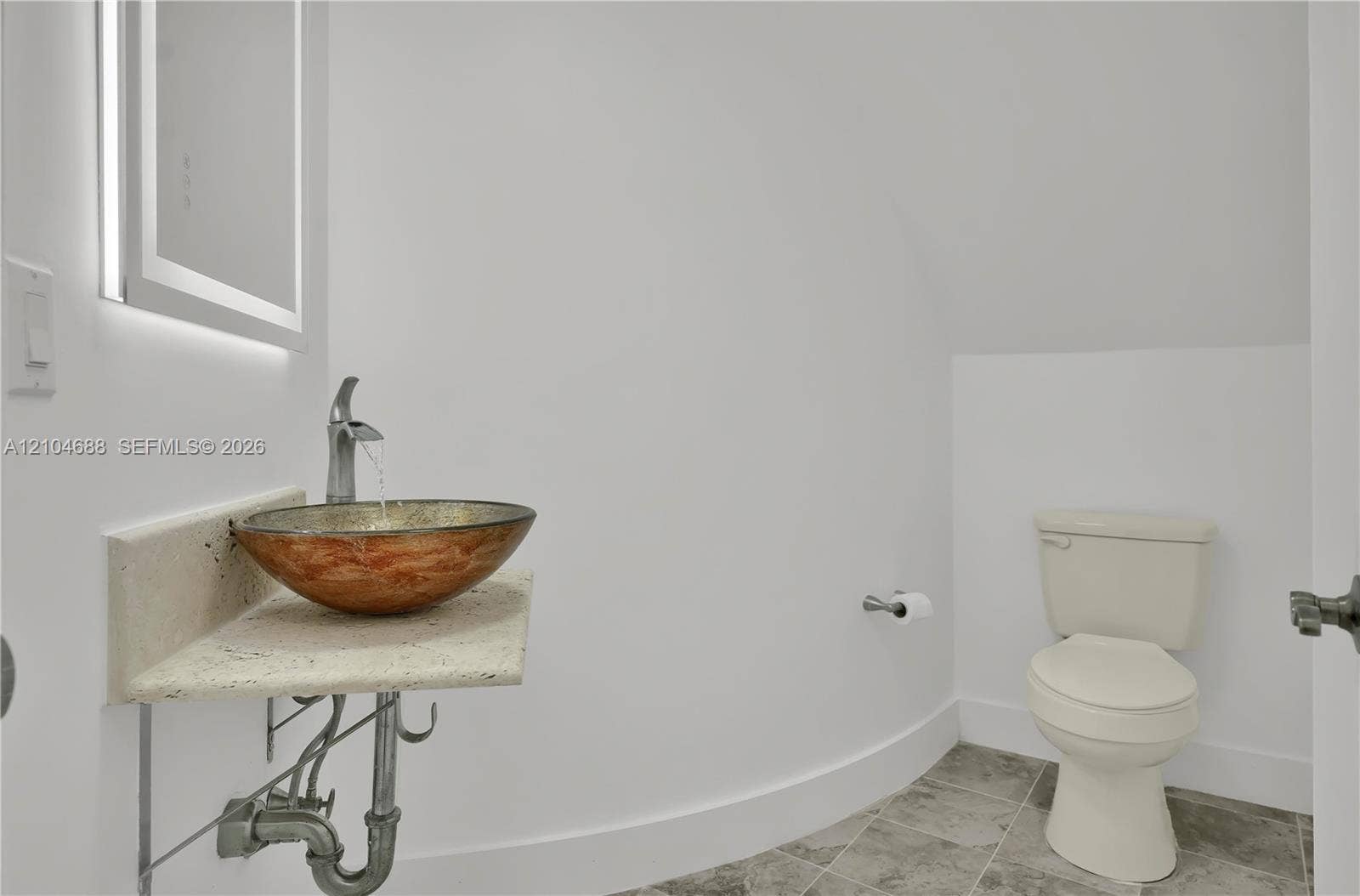 Wall-mounted floating stone vanity featuring a circular copper-tone vessel sink, brushed metal faucet, integrated LED-backlit mirror, and grey tile flooring