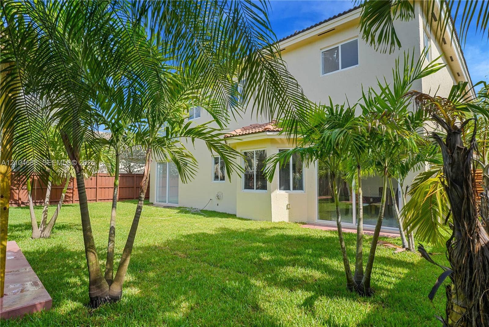 Exterior elevation featuring a stucco finish, multi-level design, sliding glass doors, a tile roof, and a fenced backyard with palm trees
