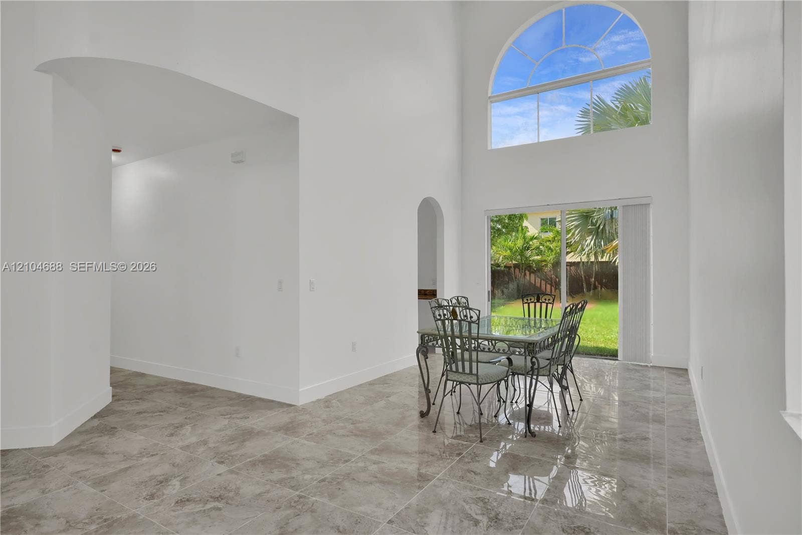 High-ceiling room featuring large-format tile flooring, arched wall openings, and a prominent floor-to-ceiling arched window with sliding glass door access to the exterior