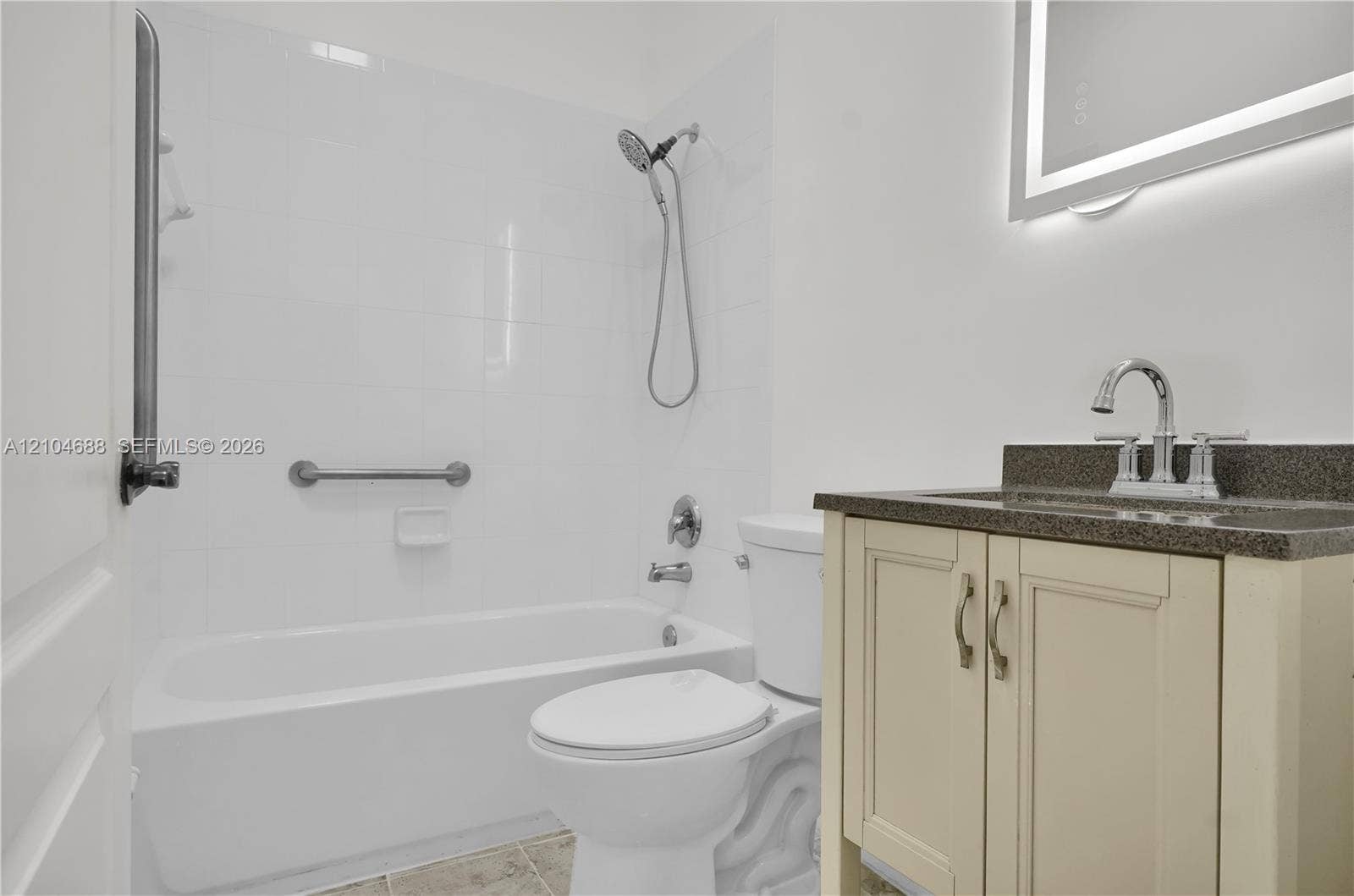 Bathroom featuring a vanity with neutral-toned cabinetry, a stone-composite countertop, an integrated sink, an illuminated vanity mirror, and a tiled tub surround with a handheld shower fixture