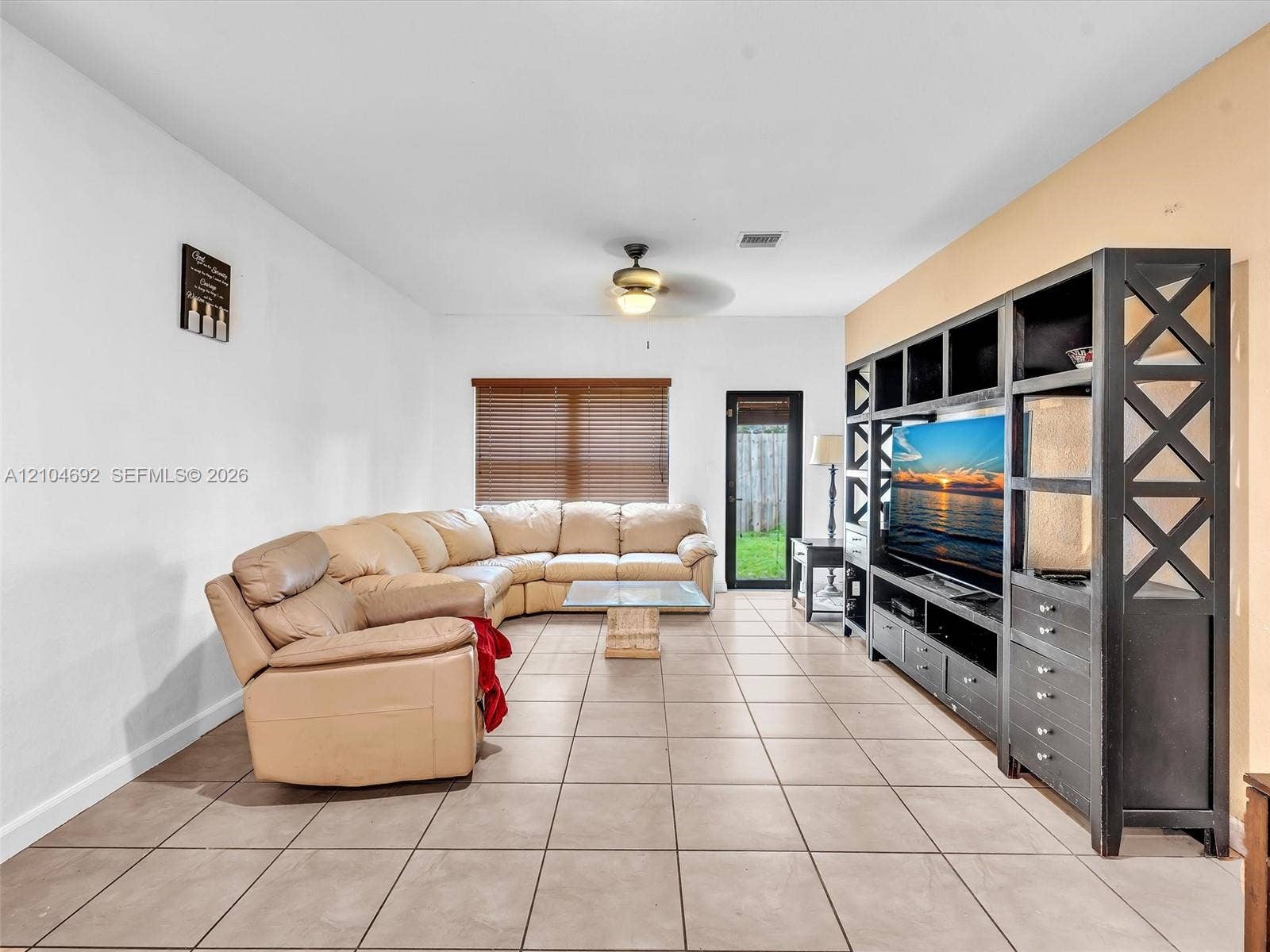 714 SE 37th Place, Homestead, FL 33033 Spacious living area featuring large-format ceramic tile flooring, built-in floor-to-ceiling wooden shelving unit, ceiling fan with integrated lighting, and horizontal window blinds