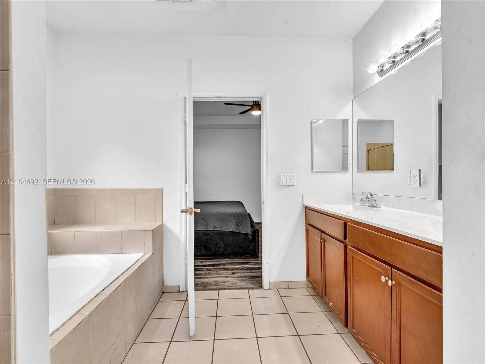 714 SE 37th Place, Homestead, FL 33033 Bathroom featuring a built-in soaking tub with neutral tile surround, a wood-finish dual vanity with a white countertop, and a full-width wall-mounted mirror