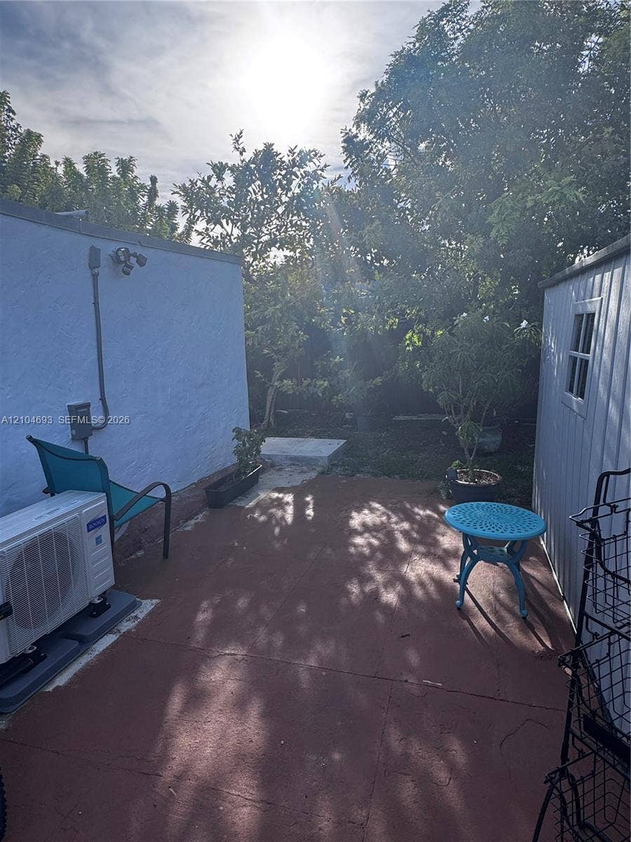 Paved patio area, white stucco exterior wall, mature trees, exterior storage shed with window, exterior lighting fixture