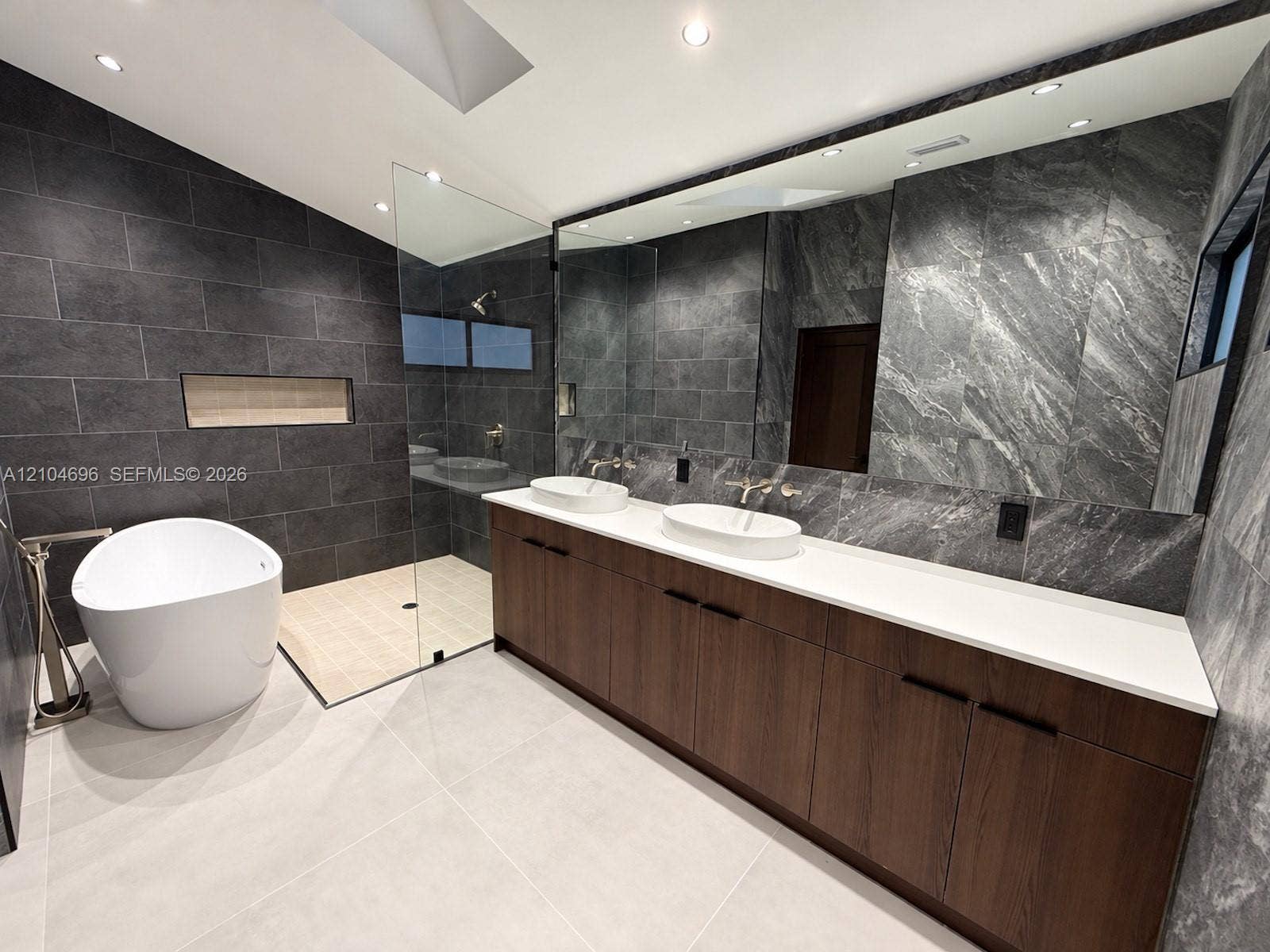 9963 NW 2nd Court, Plantation, FL 33324 Double vanity with a white solid-surface countertop and dual vessel sinks, freestanding soaking tub, walk-in shower with glass enclosure, wall-mounted matte metal fixtures, and large-format tile wall treatments
