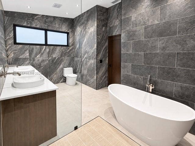 9963 NW 2nd Court, Plantation, FL 33324 Freestanding white soaking tub with wall-mounted polished chrome faucet, dual vessel sinks atop a wood-finish vanity, and floor-to-ceiling textured stone wall cladding