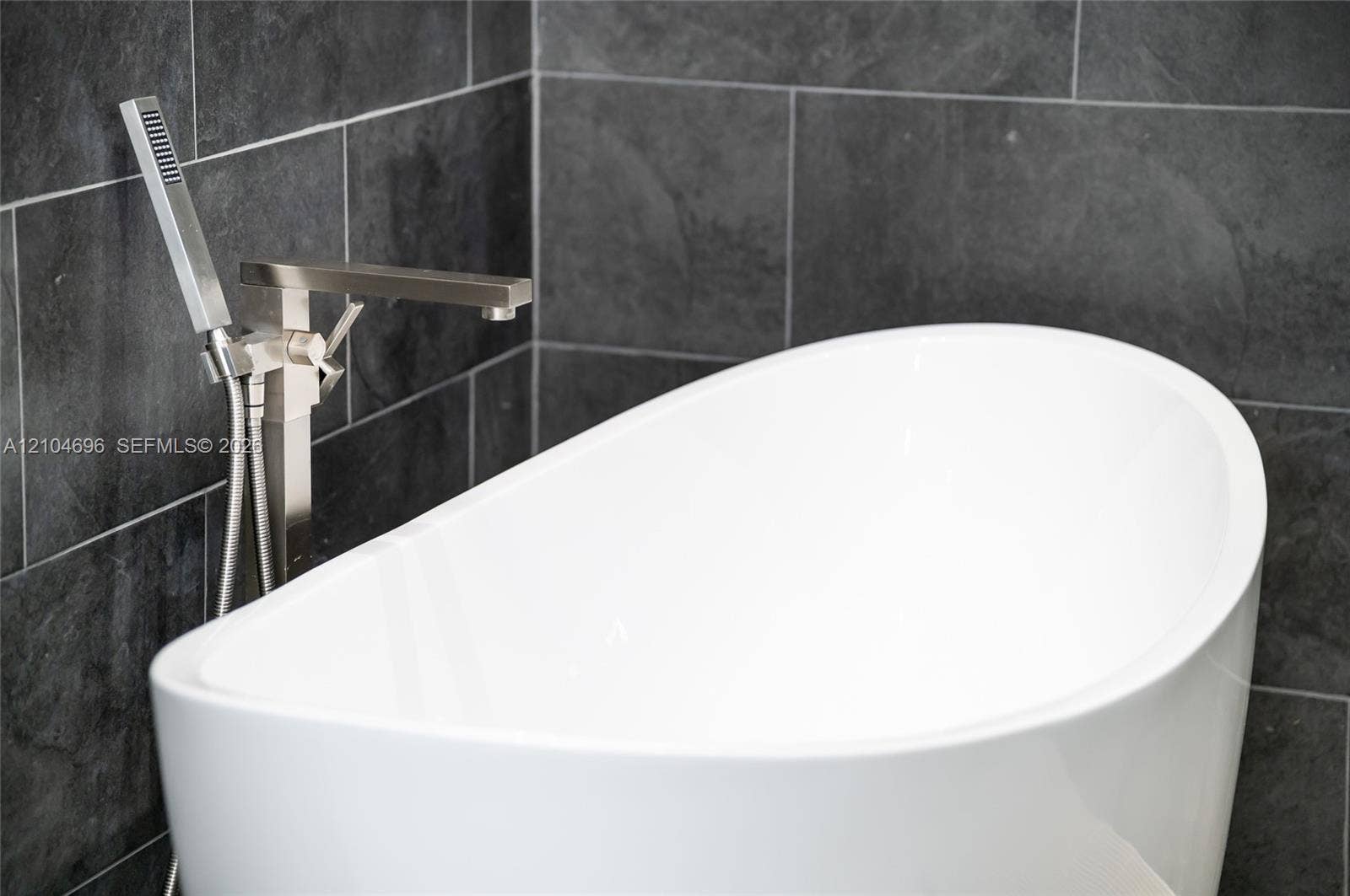 9963 NW 2nd Court, Plantation, FL 33324 Freestanding white soaking tub featuring a metallic finish floor-mounted faucet with an integrated handheld sprayer, set against charcoal-toned wall tile