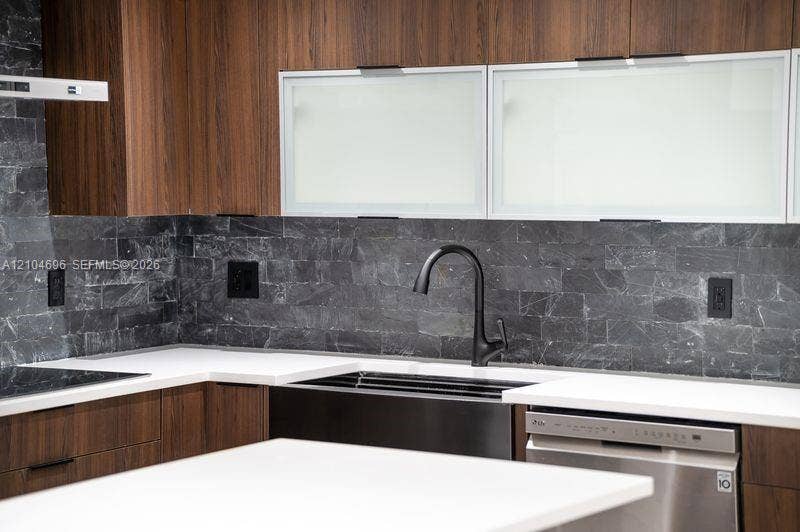 9963 NW 2nd Court, Plantation, FL 33324 Kitchen featuring wood-finish cabinetry, matte black gooseneck faucet, white solid-surface countertops, textured dark stone backsplash, and frosted glass-panel upper cabinet doors