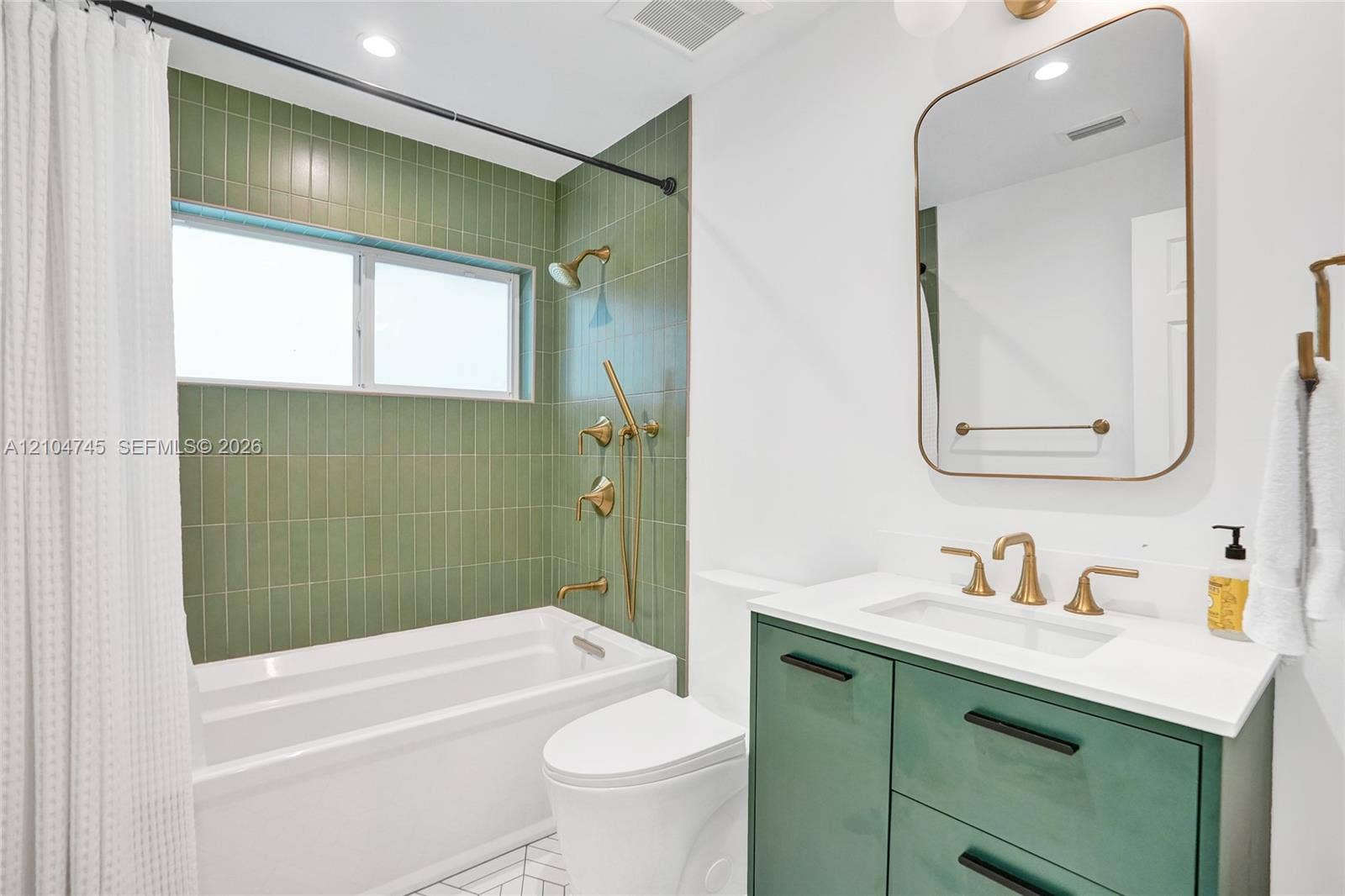 Bathtub with green vertical tile surround, gold-finish shower and bath fixtures, white vanity with green cabinetry, white countertop, and gold-finish faucet