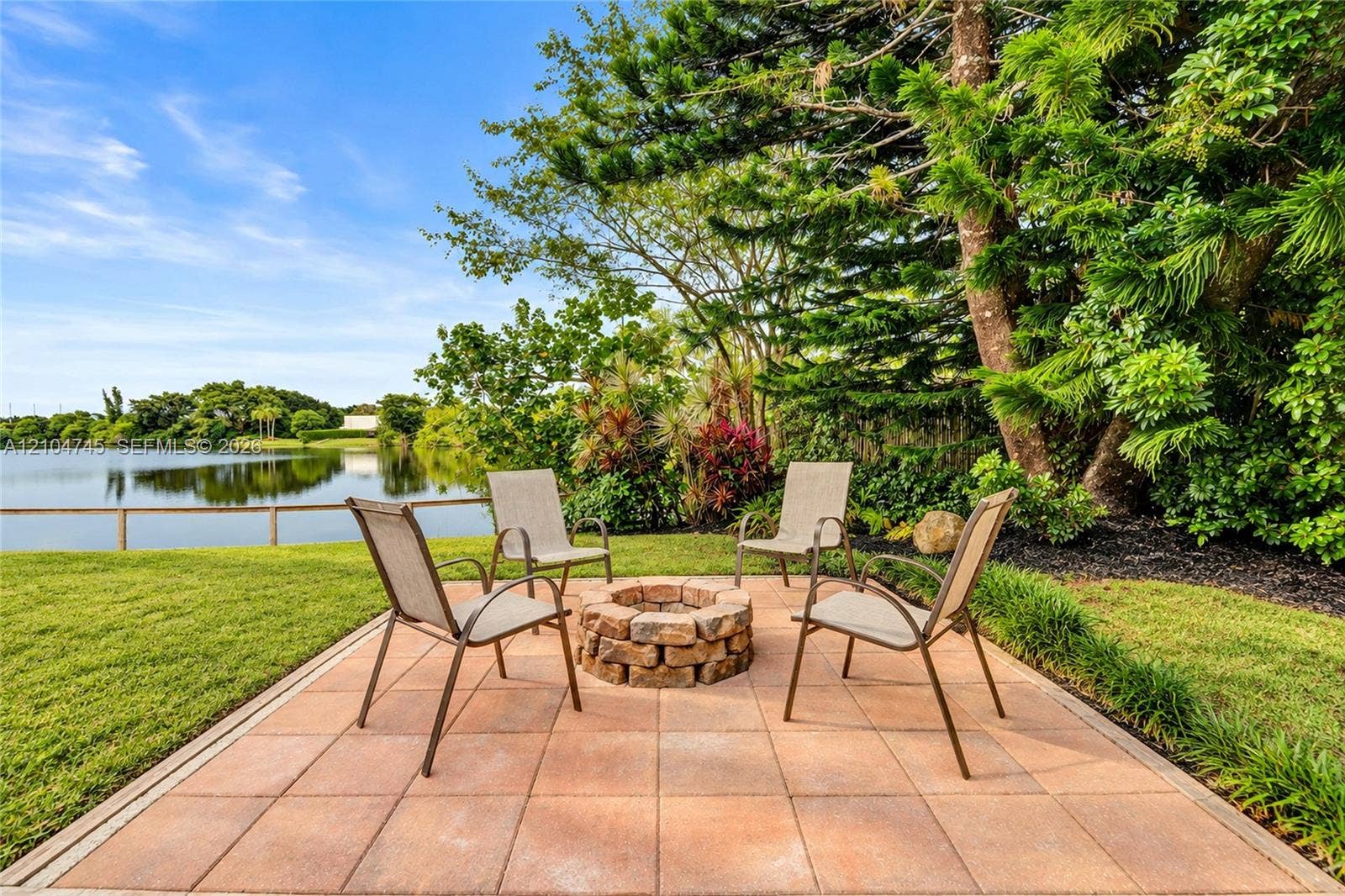 Paver patio with a stone fire pit, bordered by green lawn and diverse foliage, with a water feature in the background