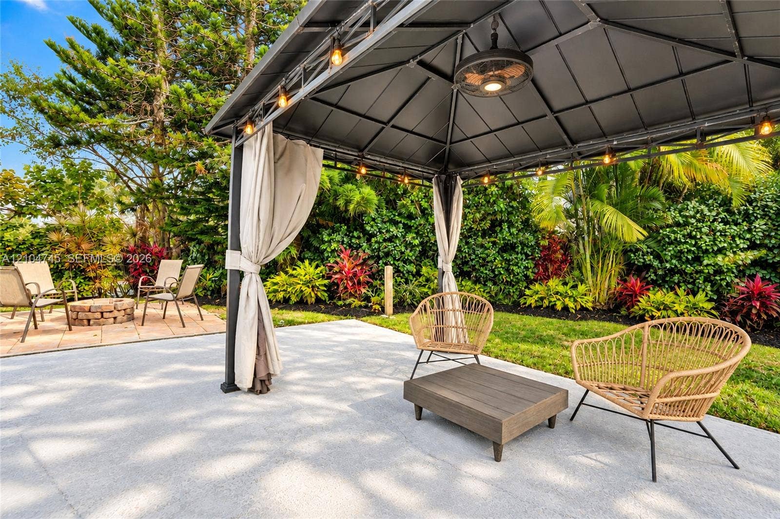 Spacious patio featuring a gazebo with integrated lighting and ceiling fan, a separate paver-stone area with a fire pit, and lush, mature landscaping