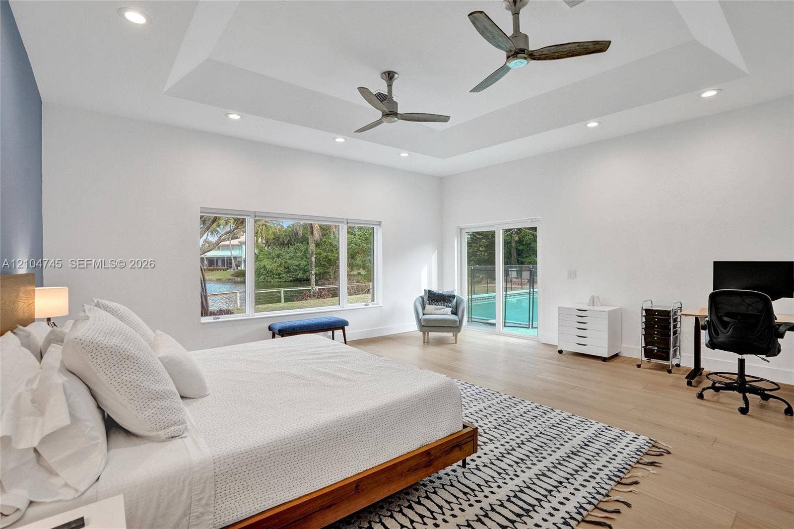 Tray ceiling with recessed lighting and two ceiling fans, wood-finish flooring, large window with water and greenery, and sliding glass door to exterior pool area