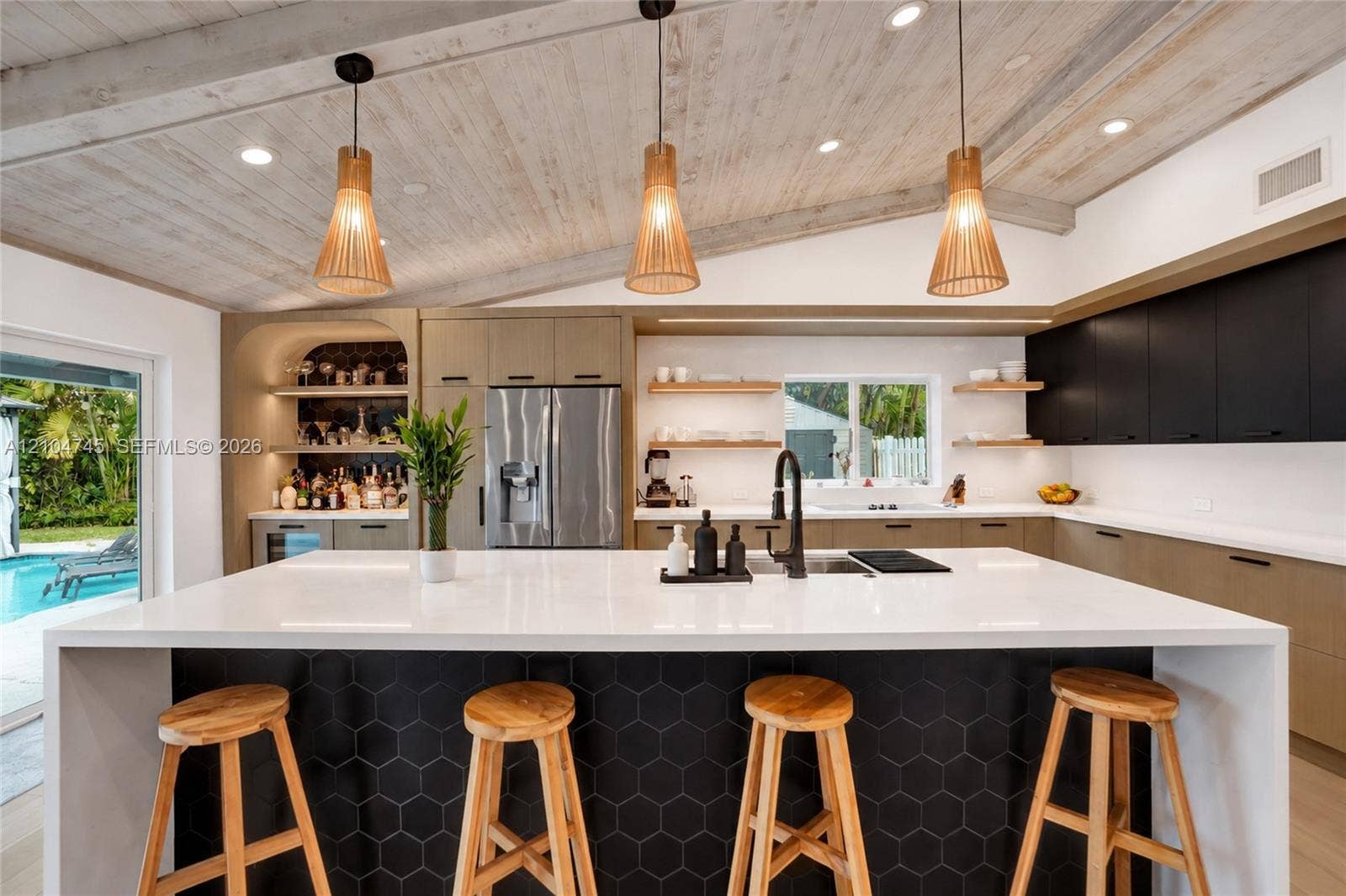 Vaulted wood-finish ceiling, large center island with hexagonal tile accent, built-in stainless steel refrigerator, integrated bar area with shelving, and direct access to an outdoor pool