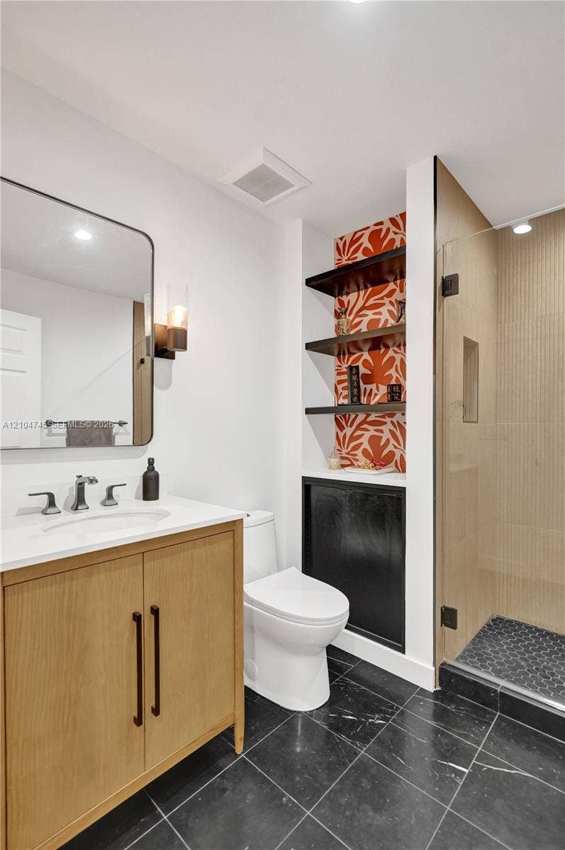 Black tile flooring, single vanity with wood-finish cabinetry, white countertop, modern wall sconce, and a glass-enclosed shower