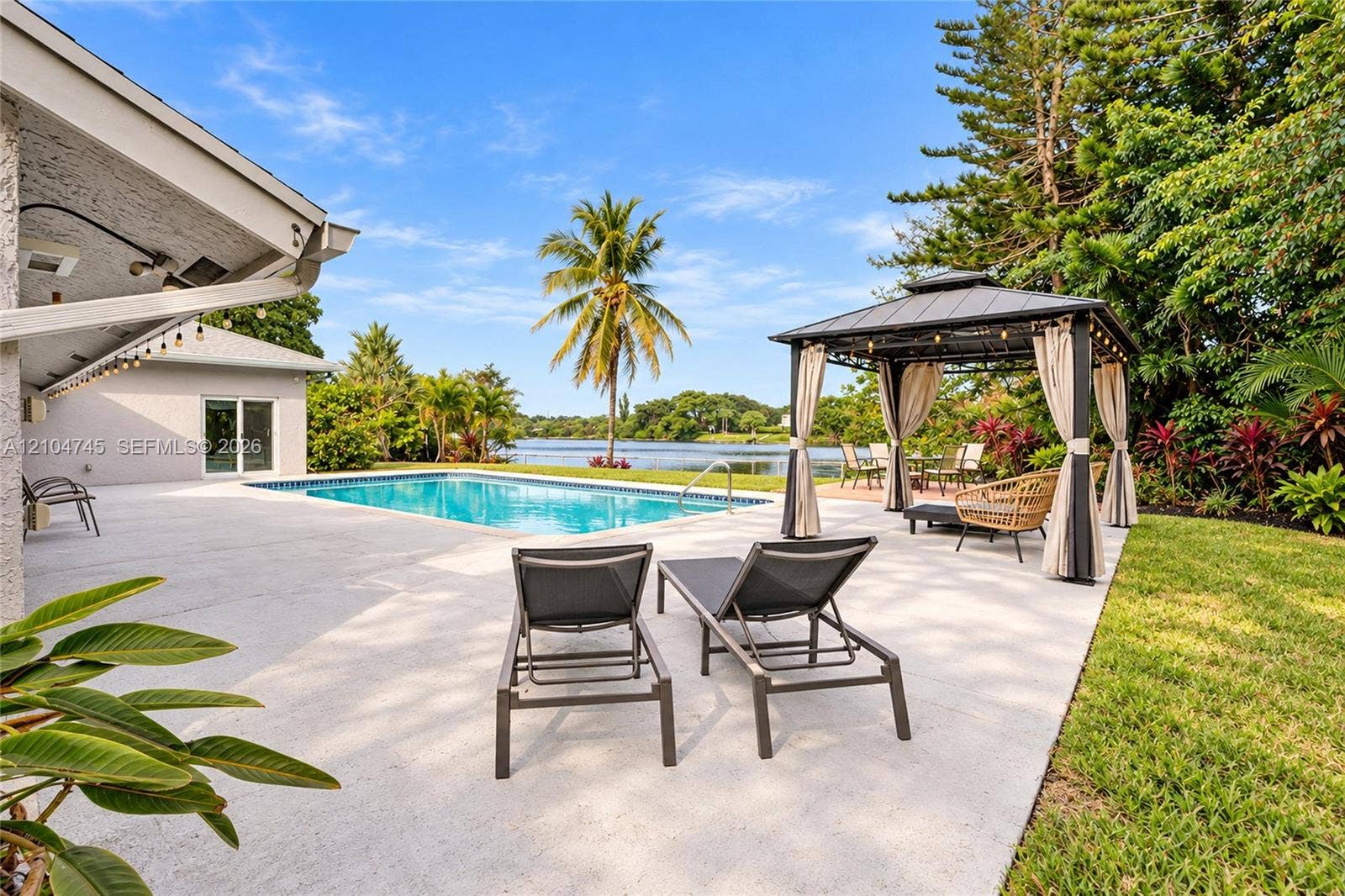 Expansive outdoor patio with a swimming pool, waterfront access, lush landscaping, and a built-in gazebo