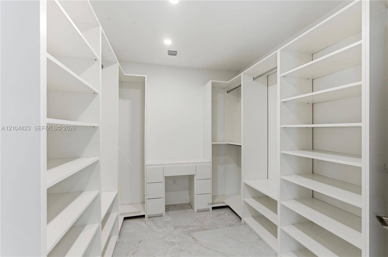 291 E Shore Drive E, Miami, FL 33133 Walk-in closet featuring extensive built-in white shelving, integrated hanging rods, a central vanity with drawers, and polished stone-finish flooring