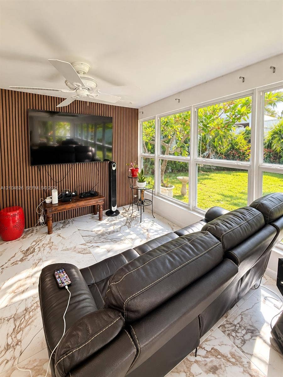 Expansive windows overlooking the exterior, white marble-look flooring, ceiling fan, and accent wall with vertical wood paneling