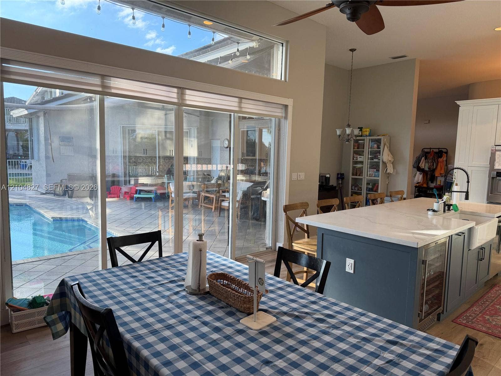 Kitchen featuring a large center island with stone-finish countertops, integrated beverage refrigerator, farmhouse sink, and shaker-style cabinetry, complemented by floor-to-ceiling sliding glass doors opening to a tiled outdoor patio and pool