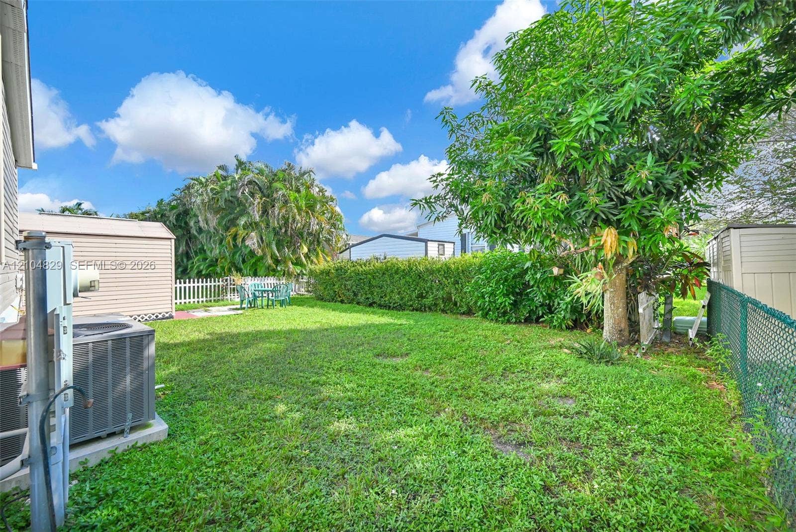 1880 SW 83rd Terrace, Davie, FL 33324 Expansive backyard with mature trees and lush lawn space, fenced perimeter, and adjacent residential structures visible in the distance under clear skies