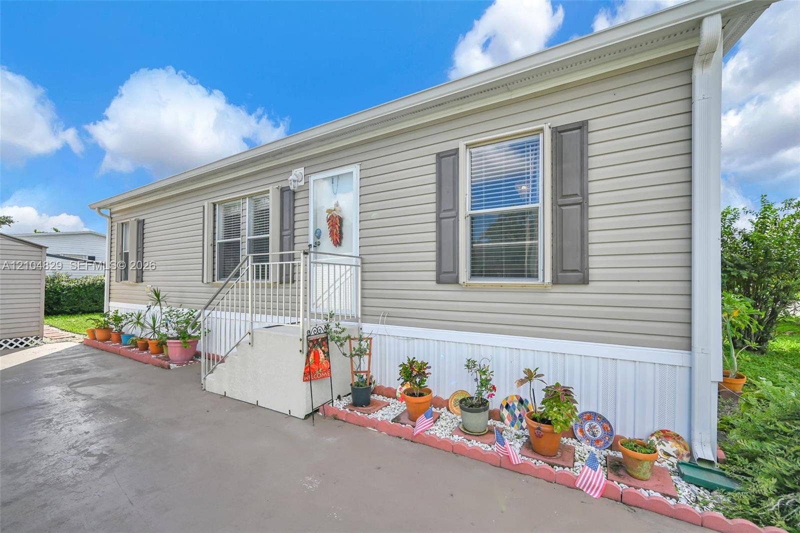 1880 SW 83rd Terrace, Davie, FL 33324 Light-colored horizontal siding exterior featuring dark-toned window shutters, a concrete driveway, a concrete landing with metal railings, and a white entrance door