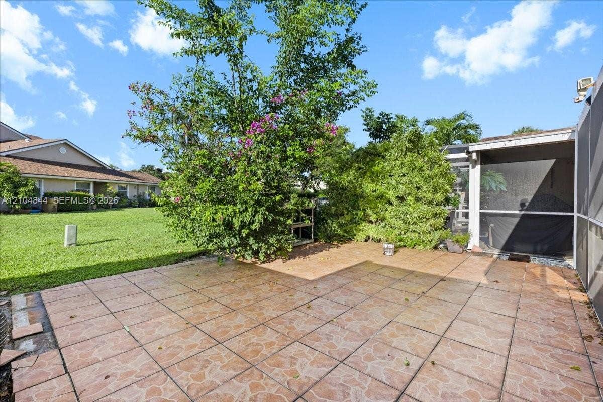 1216 NW 51st Way, Deerfield Beach, FL 33442 Rear patio featuring earth-tone ceramic tile flooring, mature flowering shrubbery, and a screened-in enclosure
