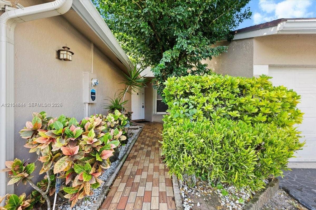 1216 NW 51st Way, Deerfield Beach, FL 33442 Entryway featuring a paver walkway, mature landscaping, stucco exterior, and wall-mounted exterior lighting