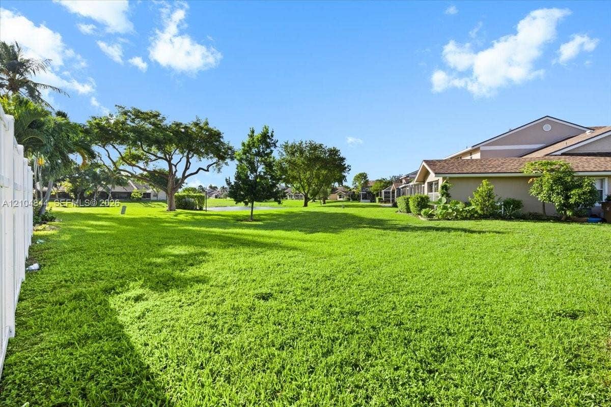 1216 NW 51st Way, Deerfield Beach, FL 33442 Expansive backyard featuring a sprawling lawn, mature trees, white vinyl privacy fencing, and exterior stucco siding with shingle roofing