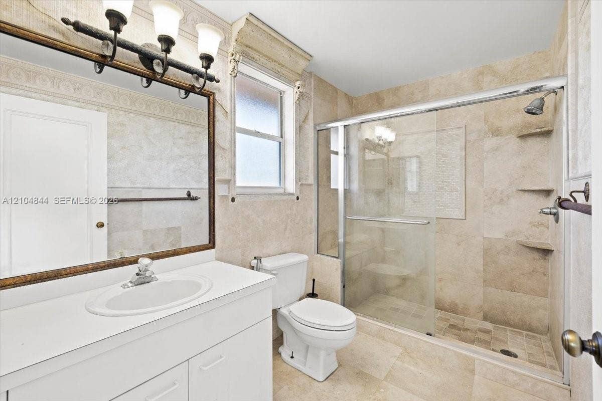 1216 NW 51st Way, Deerfield Beach, FL 33442 Bathroom featuring floor-to-ceiling stone tile, a glass-enclosed walk-in shower with integrated shelving, a white vanity with a solid-surface countertop, and a decorative crown molding accent
