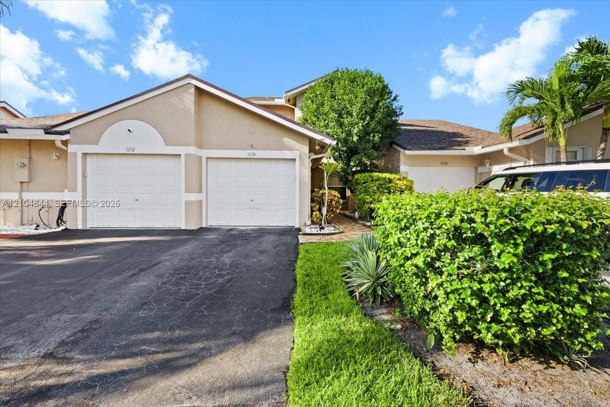 1216 NW 51st Way, Deerfield Beach, FL 33442 Stucco exterior featuring dual attached garage bays, arched architectural detailing, and landscaped grounds with mature palm trees
