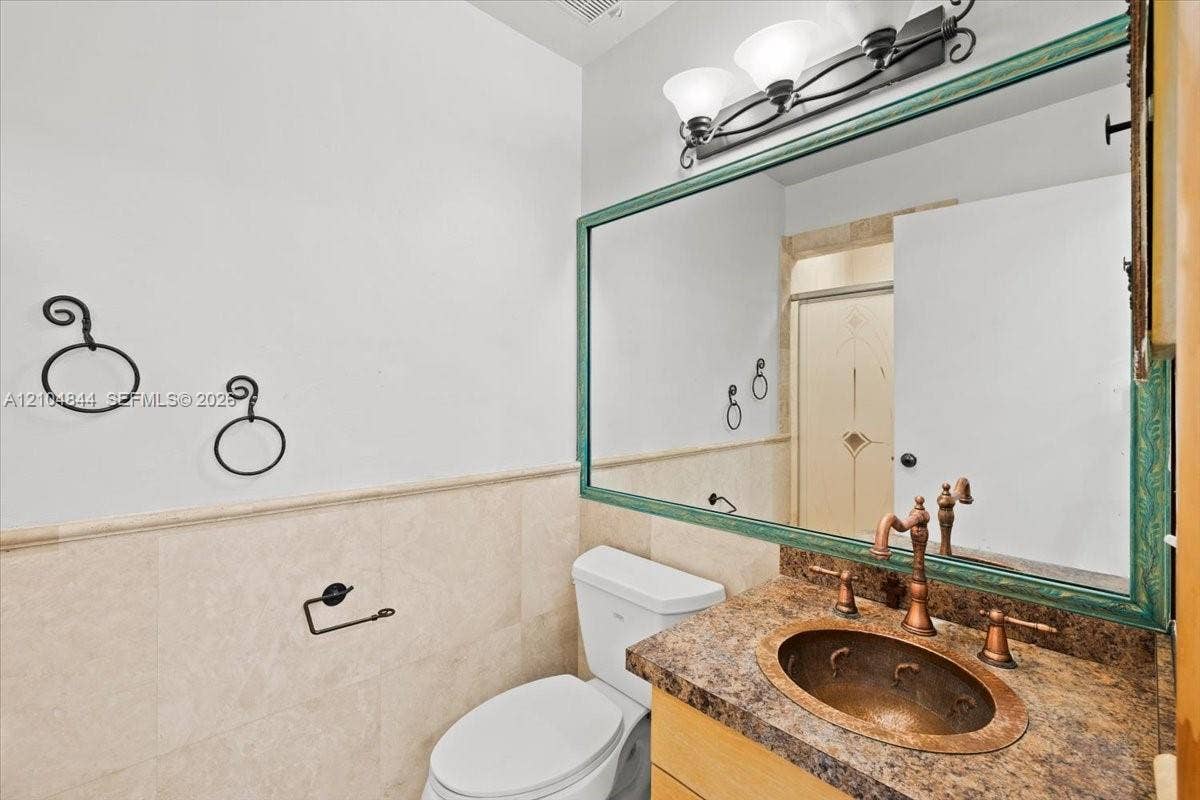 1216 NW 51st Way, Deerfield Beach, FL 33442 Bathroom vanity featuring a hammered copper drop-in sink, decorative stone countertop, wood-finish cabinetry, and a large wall-mounted mirror with a molded frame, complemented by half-height stone tile wainscoting and metallic wall-mounted hardware