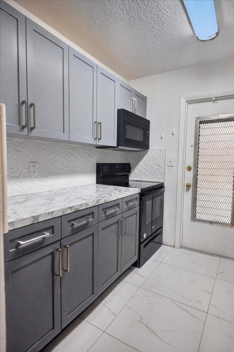 Kitchen featuring dual-tone cabinetry with modern hardware, stone-patterned countertops, textured backsplash, and large-format tile flooring