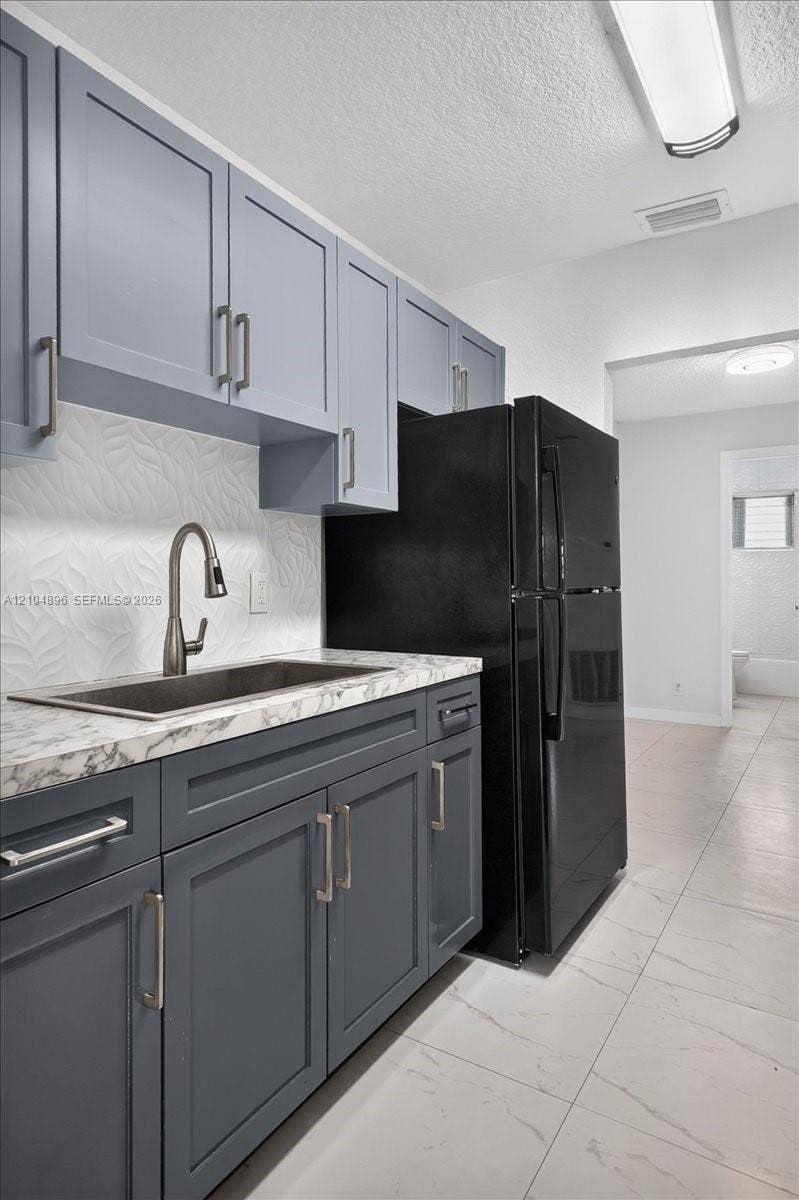 Kitchen featuring two-tone shaker-style cabinetry, stone-patterned countertops, a textured tile backsplash, an undermount sink with a gooseneck faucet, and large-format tile flooring