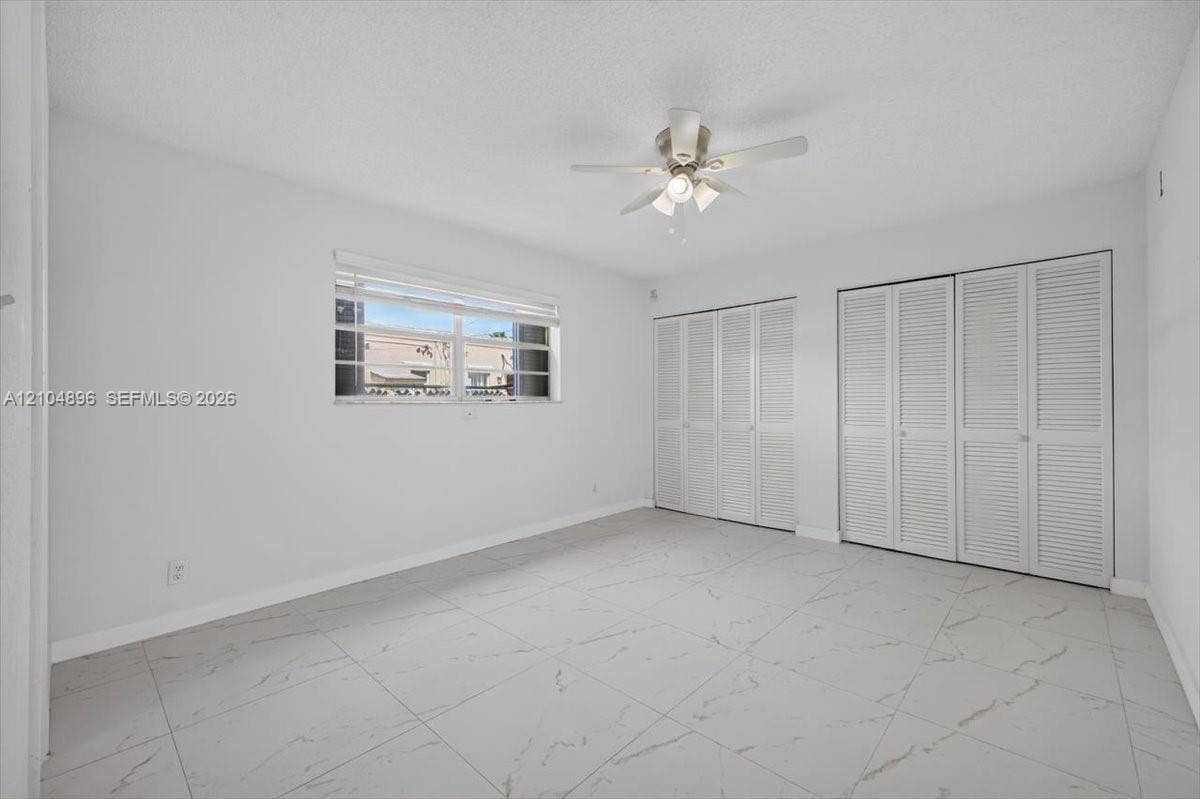 Large-format tile flooring with marble-style veining, dual sets of louvered-door closets, and a ceiling-mounted light fixture with fan