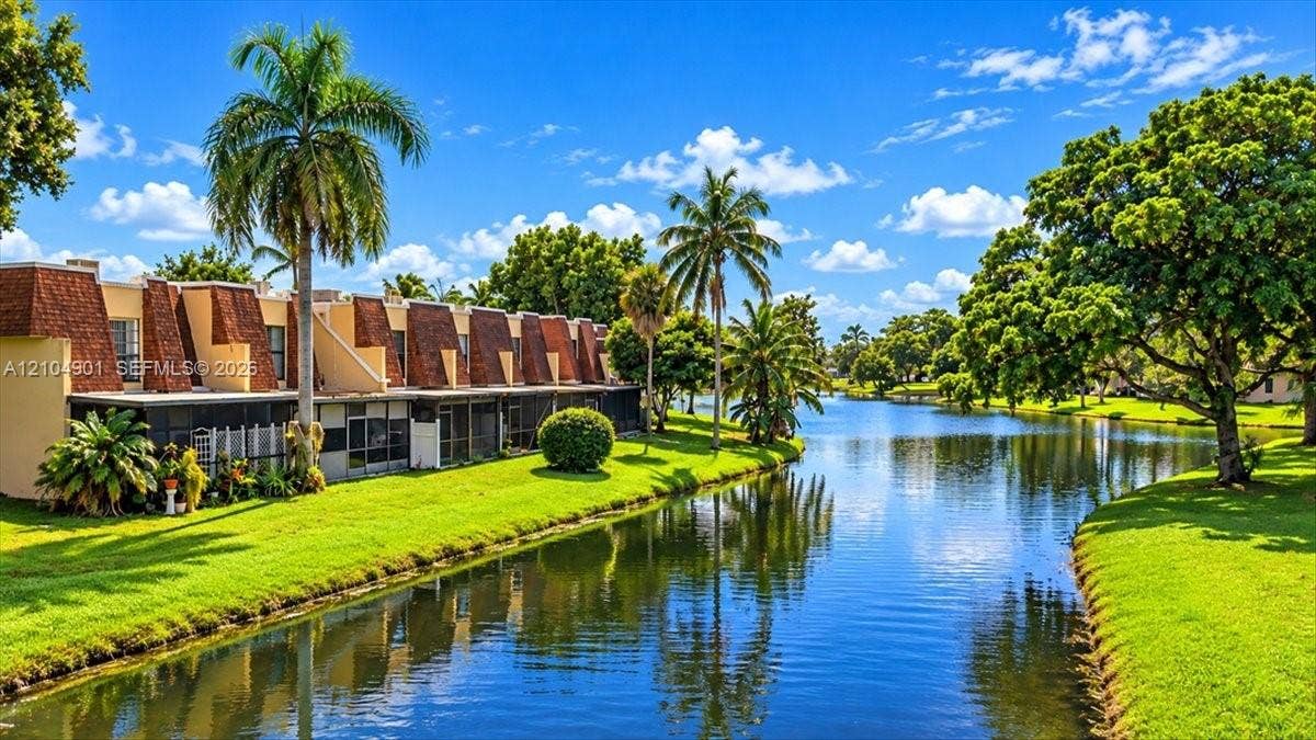 Multi-level residential exterior featuring mansard rooflines, ground-level screened enclosures, manicured lawn, and waterfront positioning