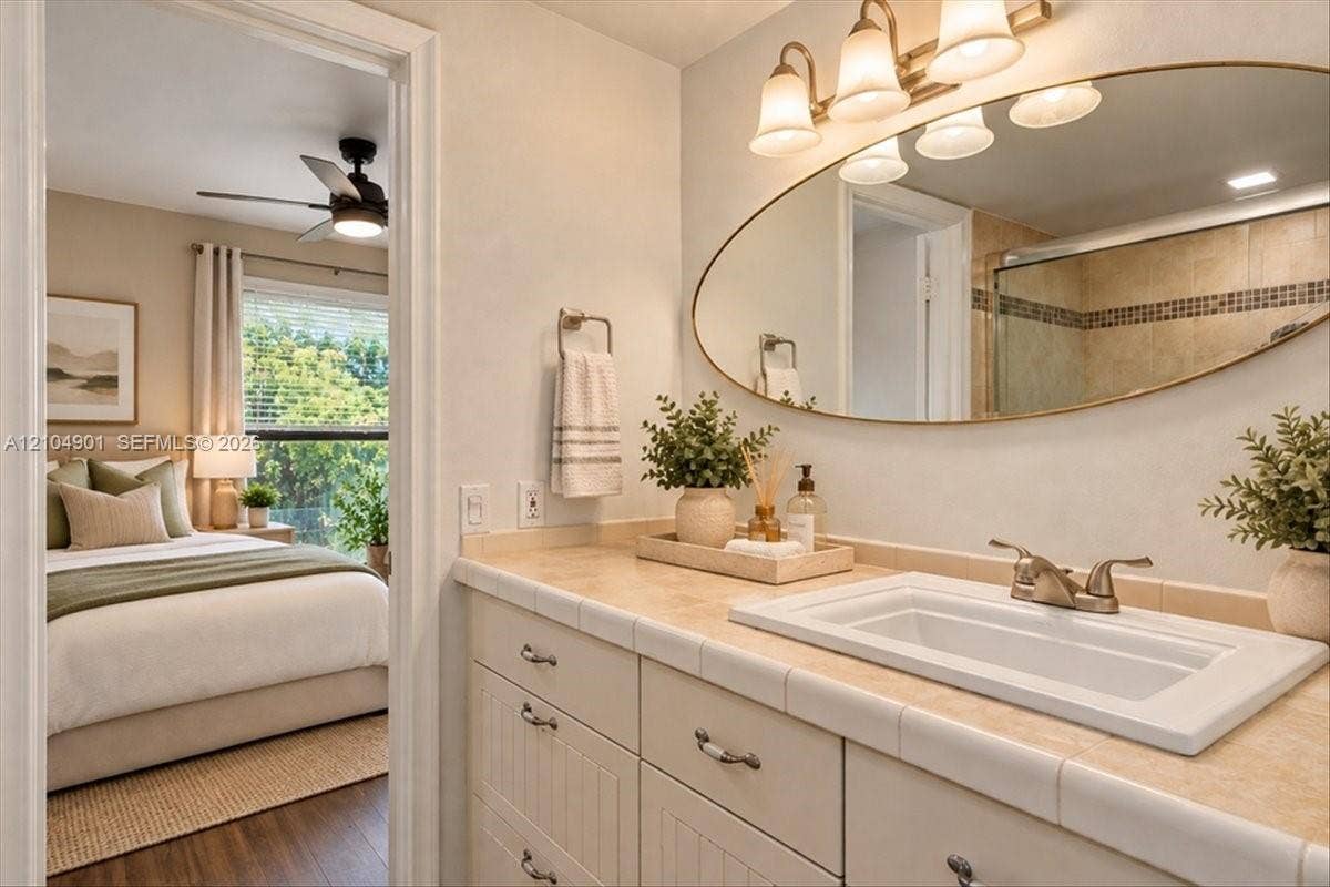 Bathroom vanity featuring a tiled countertop, drop-in sink with dual-handle faucet, oval mirror, and three-light wall fixture