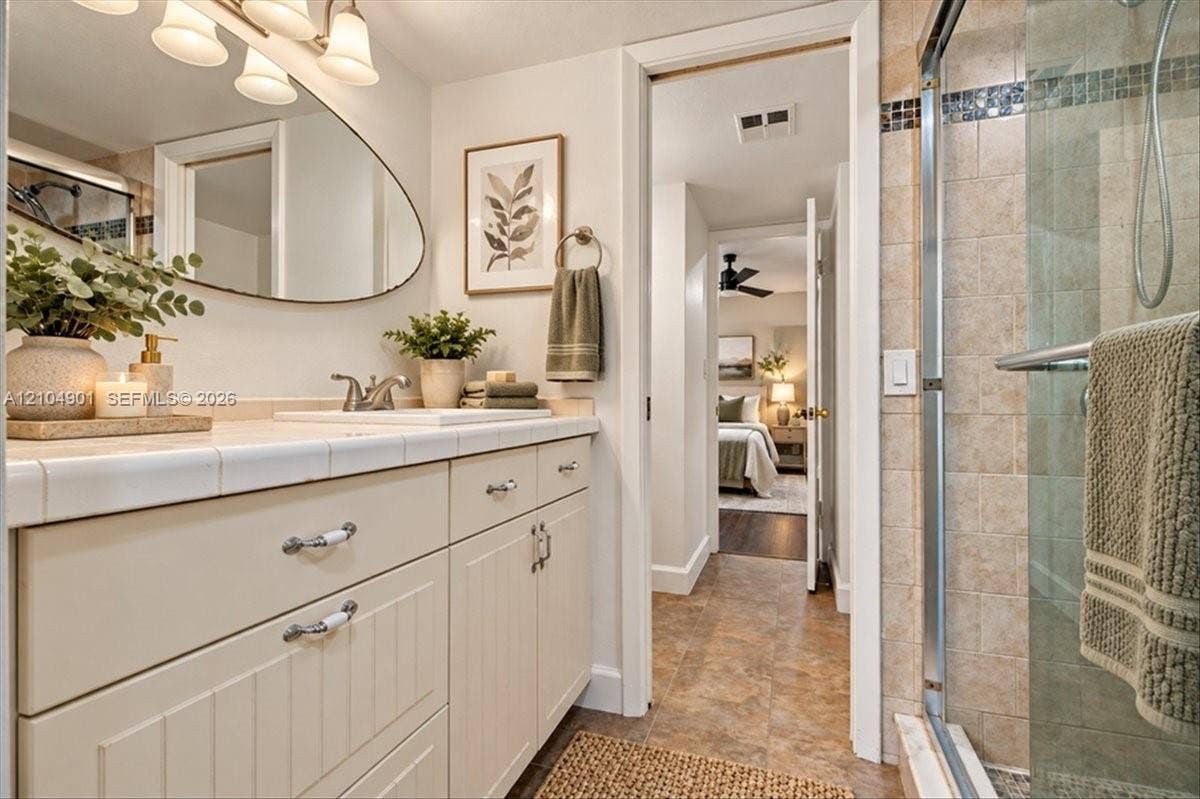 Bathroom featuring a vanity with tile-topped counter, asymmetrical wall-mounted mirror, three-light fixture, and a glass-enclosed shower with tiled walls