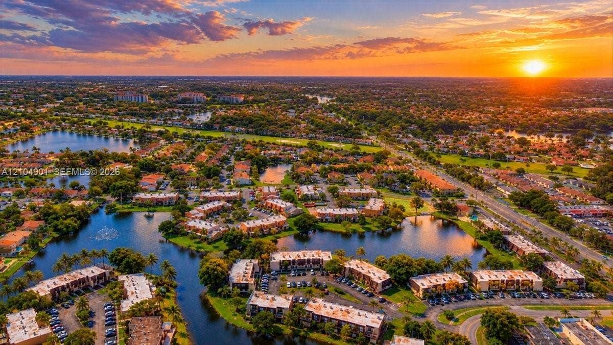 Aerial perspective of a multi-building residential complex featuring waterfront positioning, expansive inland waterways, mature palm trees, and sunset horizon