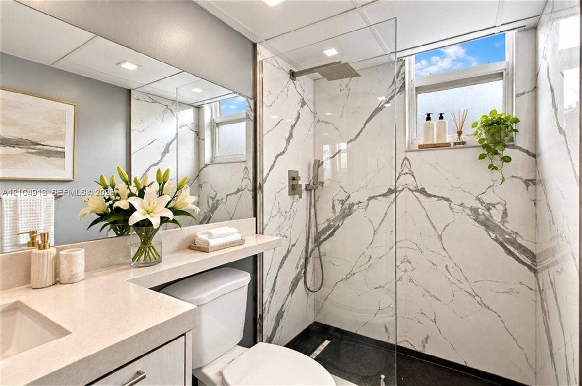 501 E Dania Beach Boulevard, #5-4A, Dania, Dania Beach, FL 33004 Bathroom featuring a walk-in shower with large-format stone-pattern tile, a frameless glass enclosure, a polished metal rainfall showerhead, and a vanity with a solid-surface countertop and full-width mirror