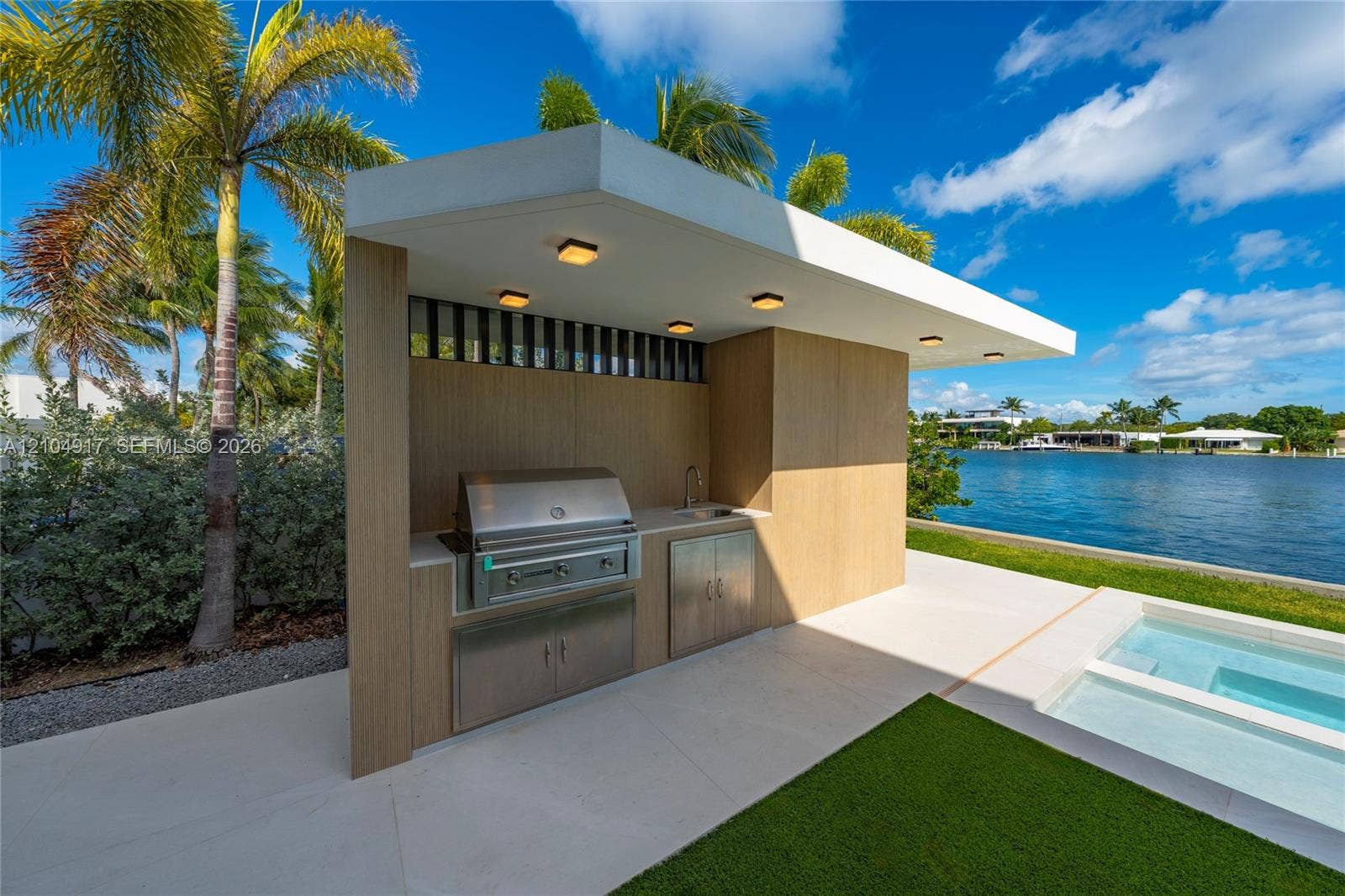 Outdoor kitchen featuring a stainless steel grill and sink, wood-finish cabinetry, recessed ceiling lighting, and a paved patio area with a built-in spa