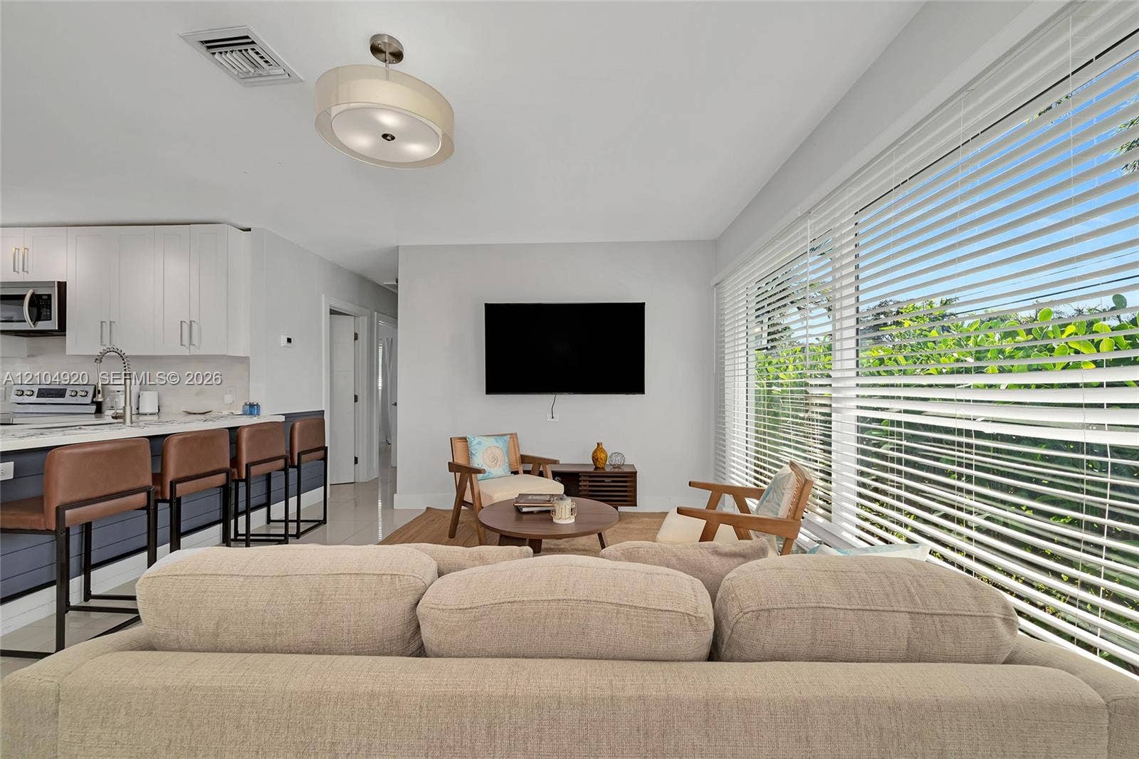 Open-concept interior featuring large-format floor tiling, custom white cabinetry, a spacious kitchen island with stone surfacing, and expansive floor-to-ceiling windows fitted with horizontal blinds