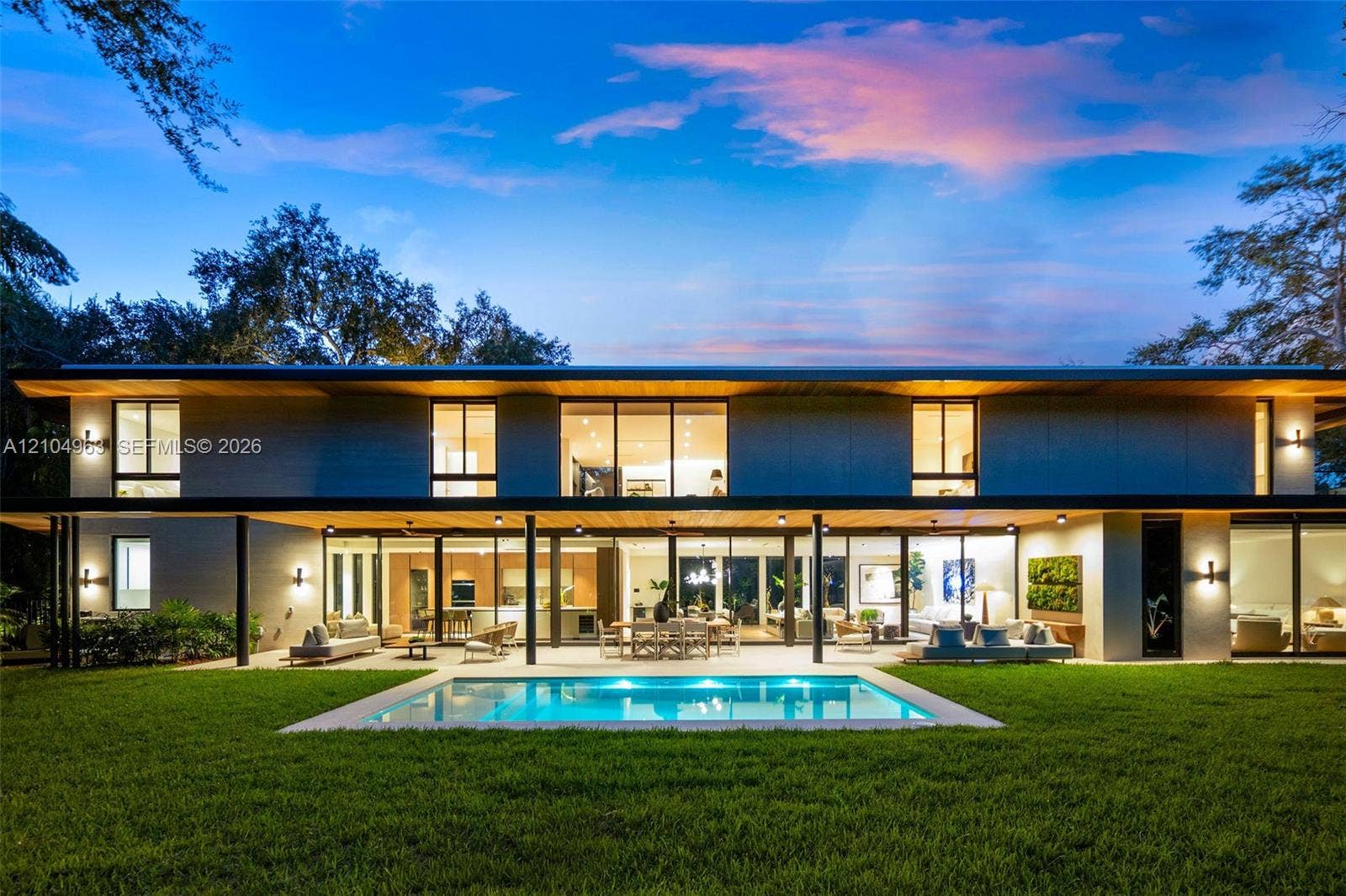 Modern two-story architectural exterior featuring expansive glass walls, a covered patio with recessed lighting, a rectangular in-ground pool, and wood-finish ceiling accents