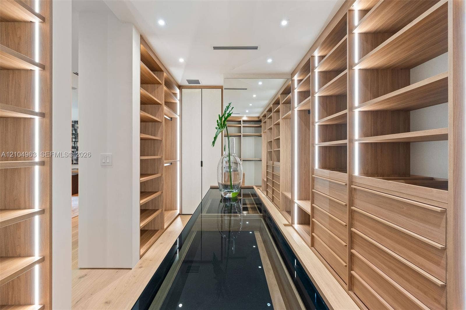 Custom storage cabinetry featuring light wood-finish shelving, integrated vertical LED lighting, a glass-topped center island, and recessed ceiling lighting